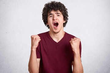 Indoor shot of successful happy student opening mouth widely, shouting enthusiastically, raising arms, having clenched fists, being in high spirits, standing isolated over white background in studio.