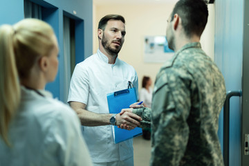 Mid adult doctor shaking hands with military officer at hospital hallway.