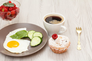 Plate with fried egg, fresh cut cucumber and leaf of basil, fork, cup of black coffee, muffin and glass bowl with fresh raspberries