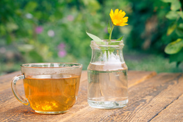 Calendula flower with a stem in a glass jar and cup of green tea.