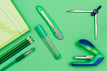 Book, color pencils, pen, felt-tip pen, paper knife, compass and stapler on green background.