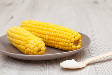 Porcelain plate with boiled corn and wooden spoon with salt on a table.