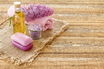 Bottle with oil, bowl with sea salt, soap and towel with lilac flowers on wooden background.
