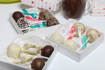 Cake pops decorated with a bow of braid, packed in a gift box. Nearby are cups of melted chocolate.