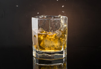 A glass of whiskey with ice on a dark color photo with splashes from falling ice