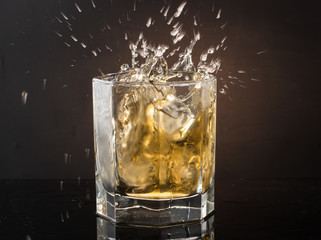 A glass of whiskey with ice on a dark color photo with splashes from falling ice