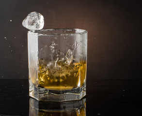 A glass of whiskey with ice on a dark color photo with splashes from falling ice