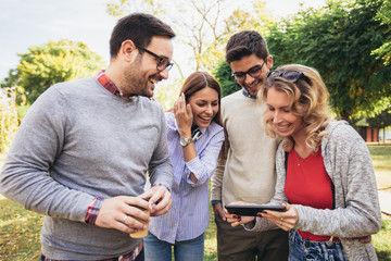 Four happy smiling young friends walking outdoors in the park holding digital tablet