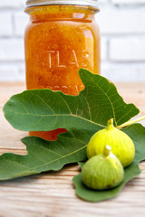 Antique glass preserve bottle with fig preserve together with a fresh green fig leaf and halved ripe figs on a raw wood surface. 