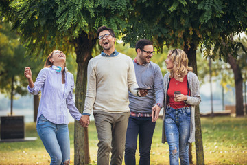 Four happy smiling young friends walking outdoors in the park holding digital tablet