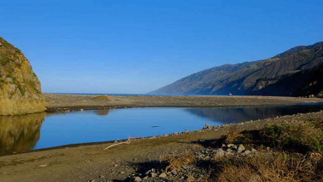 Slow pan of coastal lagoon on calm morning stillness, sand berm and large imposing Big Sur coastline mountains in background