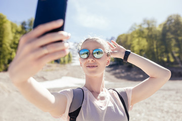 Naklejka premium Beautiful young girl traveler environmentalist makes selfie in the mountains during the sunny summer weather. Concept candid lifestyle and trekking in the mountains
