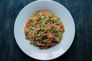 Asian cuisine. Salad of cellophane noodles, fried with vegetables, decorated with greens and crab sticks. Funchoza. Proper nutrition. Healthy food. View from above. Dark wooden background.