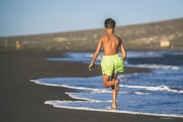 Fototapeta na wymiar Back view of young boy running on the beach after the end of school Student enjoying summer vacation playing on the seashore Teen runs happy with a warm evening light Concept of freedom and happiness