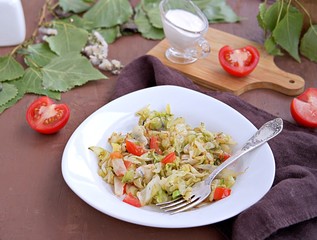 Fried cabbage with tomatoes in a white plate on a dark brown background. Served with sour cream.
