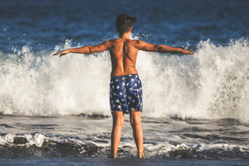 Back view of a young boy playing happy at the beach after the end of school. Child waiting waves on the seashore. Student enjoying holidays and summer vacation Concept of happiness and freedom