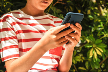 Close up view of a boy playing with smartphone outdoor. Closeup image of young hands writing a message on phone. New technology allow teen to communicate with remote friends. Child make a video call