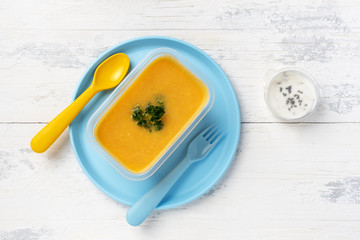 vegetable lentil soup in a plastic box on a plastic colored plate with plastic spoon and fork. on a white wooden table. with box with sour cream