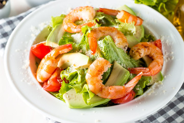Fresh shrimp salad made of tomato, ruccola, avocado, prawns, chicken breast, arugula, crackers and spices. Caesar salad in a white, transparent bowl on wooden background