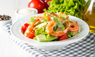 Fresh shrimp salad made of tomato, ruccola, avocado, prawns, chicken breast, arugula, crackers and spices. Caesar salad in a white, transparent bowl on wooden background