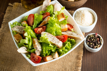Fresh salad made of tomato, ruccola, chicken breast, eggs, arugula, crackers and spices. Caesar salad in a white, transparent bowl on wooden background