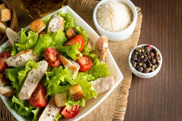 Fresh salad made of tomato, ruccola, chicken breast, eggs, arugula, crackers and spices. Caesar salad in a white, transparent bowl on wooden background