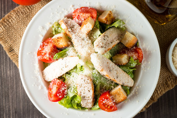 Fresh salad made of tomato, ruccola, chicken breast, eggs, arugula, crackers and spices. Caesar salad in a white, transparent bowl on wooden background