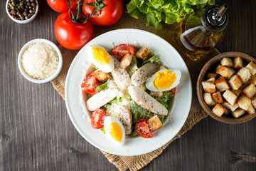 Fresh salad made of tomato, ruccola, chicken breast, eggs, arugula, crackers and spices. Caesar salad in a white, transparent bowl on wooden background