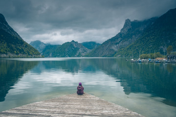 Kobieta samotnie siedzi na drewnianym pomoście nad górskim jeziorem Traunsee w Austrii w pochmurny dzień	