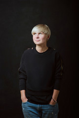 Portrait of stylish blonde short hair young woman in black sweatshirt and jeans sitting on dark background 