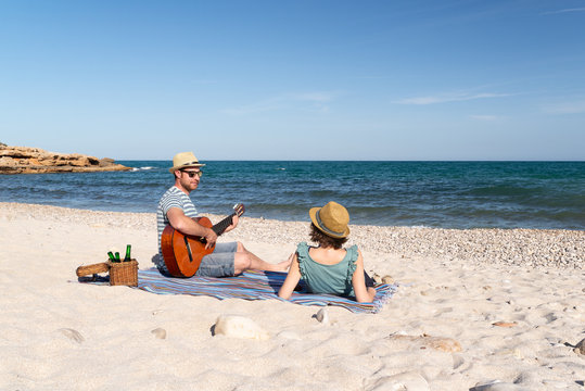 Couple in love having a picnic and playing the guitar at a mediterranean beach at sunset. Romantic weekend