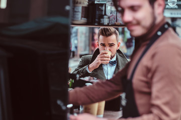 Handsome groomed man is enjoying his coffee while sitting at coffeeteria. There are working barista at frontground.