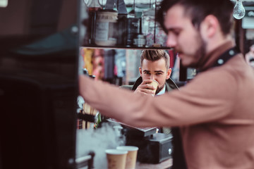 Handsome groomed man is enjoying his coffee while sitting at coffeeteria. There are working barista at frontground.