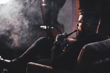 Groomed bearded man is relaxing on lounge near fireplace while smoking hookah. He has tattoo on his...