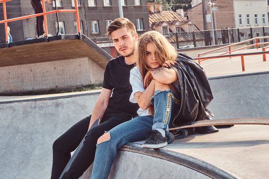 Young attractive couple of students are sitting at skatepark with their longboards.