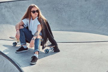 Trendy young woman with long hair and sunglasses is sitting at skatepark on her longboard.