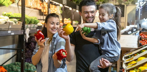 Fototapeta premium Happy family buys vegetables. Cheerful family of three choosing tomatoes in vegetable department of supermarket or market.