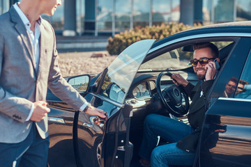 Handsome businessman in sunglasses is talking by mobile phone while his young smart assistant is opening door for him.