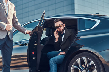 Attractive serious man in sunglasses is talking by smartphone and sitting in the car while his...
