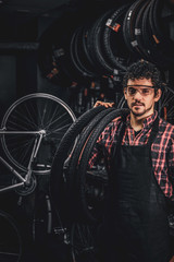 Fototapeta na wymiar Pensive happy man in protective glasses is standing with tires at bicycle repairing studio.
