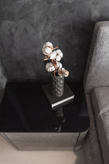 Dried cotton branch in stylish ceramic vase stands on black book and modern gret bedside table. Interior details.