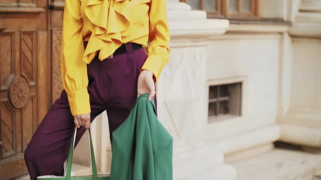 Street fashion full body portrait of young beautiful elegant lady wearing trendy color sunglasses, headband, stylish yellow blouse, purple trousers, holding green bag, posing in old european city