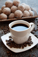 Black coffee in a white cup next to a tray with mini donuts sprinkled with powdered sugar