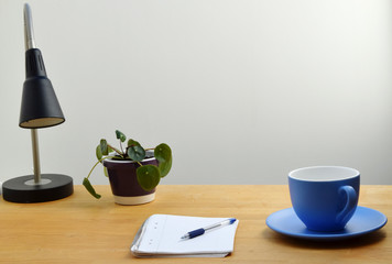 Study desk with notepad, pen, lamp, chinese money plant (pilea peperomioides) in a pot, and large cup of coffee on wooden desk
