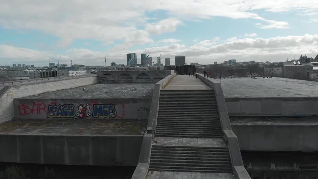 Large gray architectural complex or bridge with two staircases and walking people against blue cloudy sky and city buildings in early spring. Stock. Urban architecture