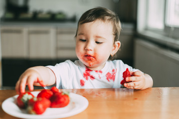Little girl child in white t-shirt eating strawberries all smeared and dirty
