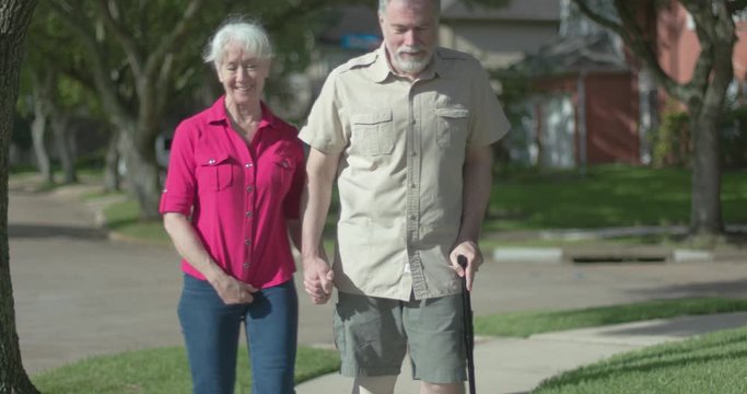 A mature woman accompanies her husband as he walks slowly with his cane in an effort to rehabilitate his knee after surgery.