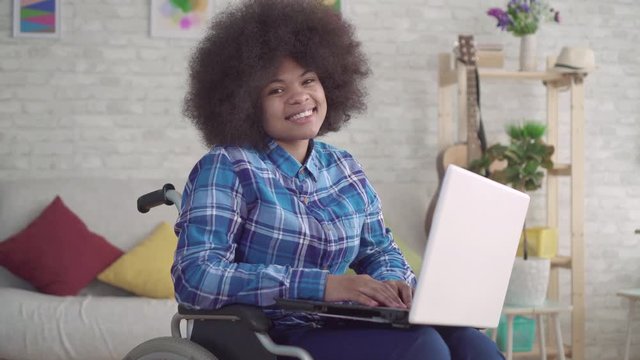 disabled african american woman with an afro hairstyle in a wheelchair uses a laptop looking at the camera