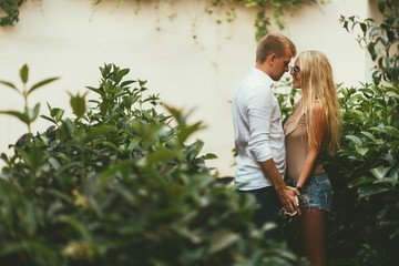 Stylish young couple on a date stands in beautiful tropical greenery and gently holding hands. The girl with long hair and in sunglasses smiles a handsome guy in a white shirt. Romance, relationship