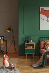 Real photo of a kids room interior with a black lamp between a desk with laptop and bed with green...